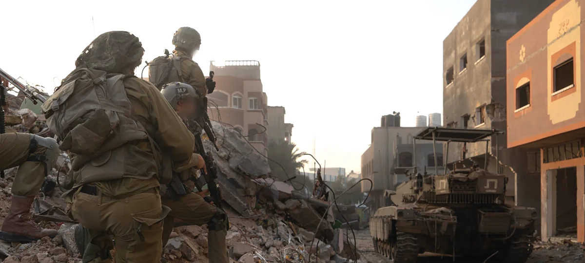 Soldiers crouch on a pile of rubble beside an armored vehicle in a war-damaged street