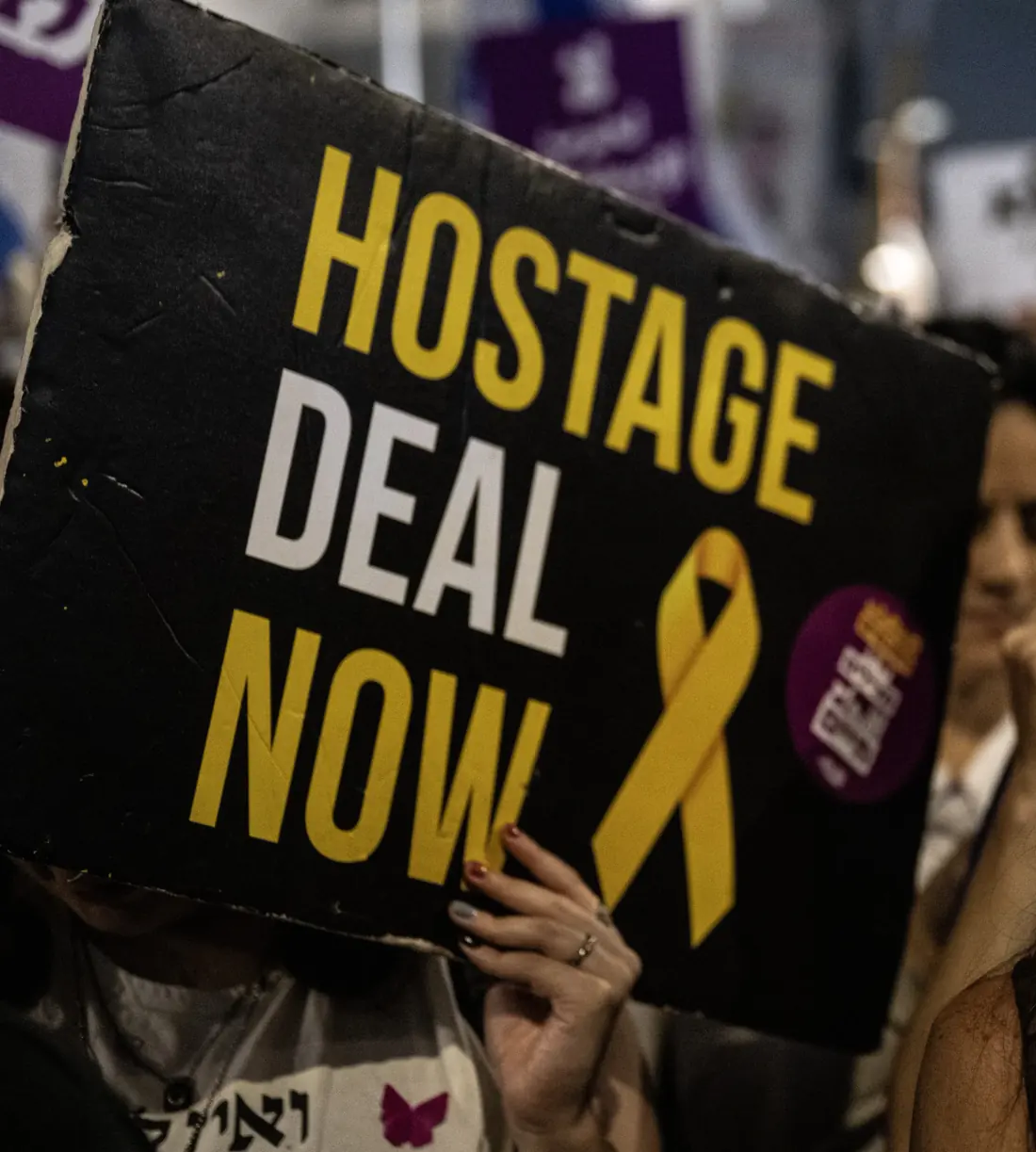 A protester holds a black sign reading "HOSTAGE DEAL NOW" with a yellow ribbon at an evening rally