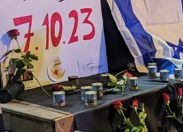 A memorial table with memorial candles and red roses beside a "7.10.23" sign and an Israeli flag