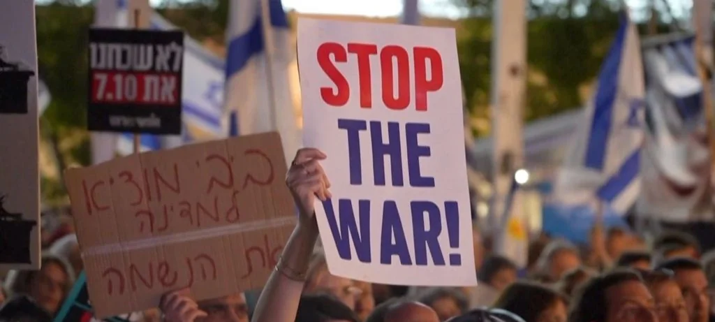 Protesters hold Hebrew and English signs, including one reading "STOP THE WAR!", among Israeli flags