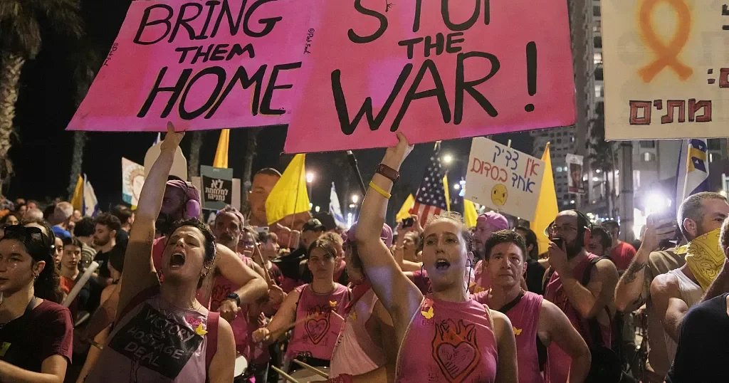 Women in pink shirts chant at a night protest under signs reading "BRING THEM HOME" and "STOP THE WAR!"