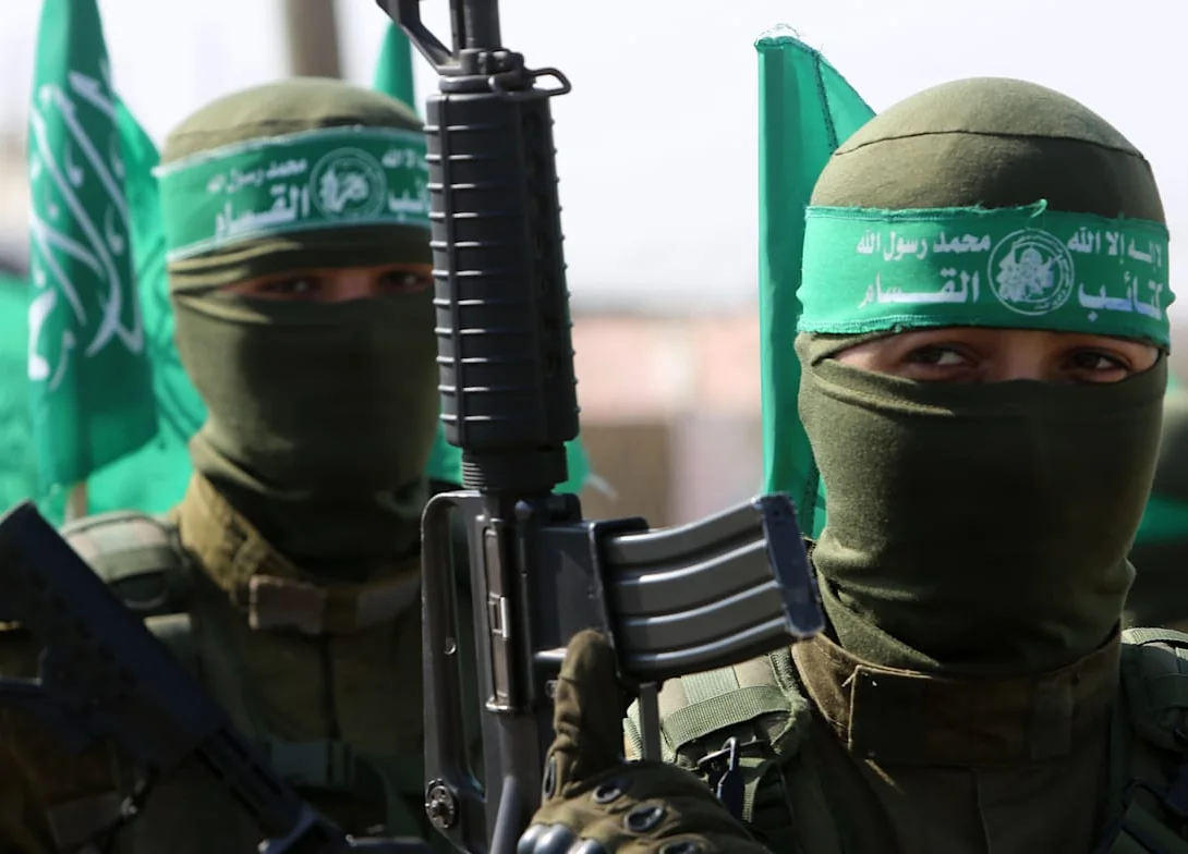 Two masked gunmen wearing green Qassam Brigades headbands, one holding a rifle, with green Hamas flags behind them