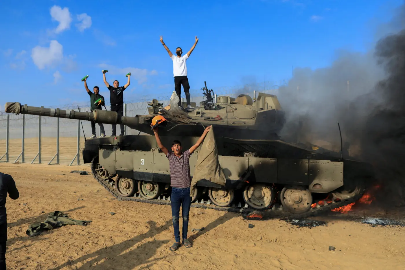 Men celebrating on and beside a burning military tank near the border fence, black smoke rising from it