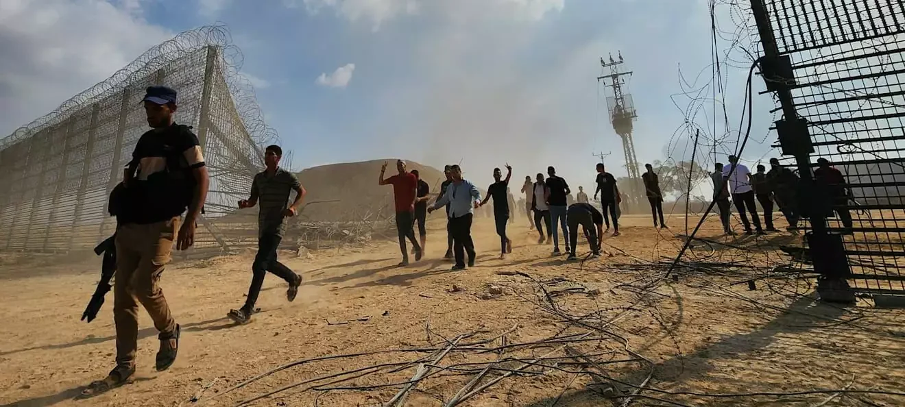 People cross on foot through a breached border fence in sandy terrain, one of them carrying a rifle