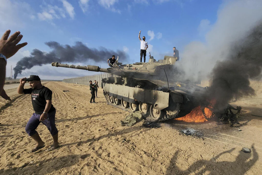 Men stand on a burning military tank in sandy ground near the border fence as black smoke rises